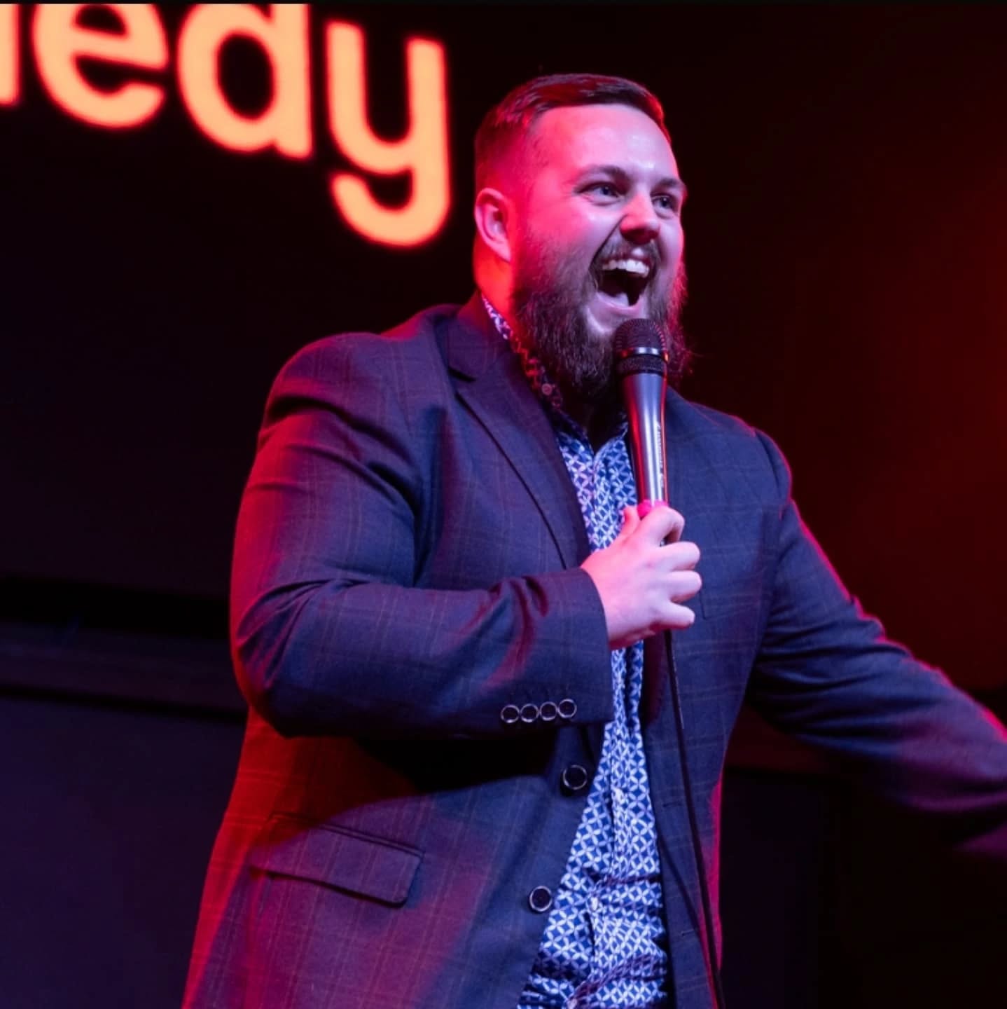 Jamie on stage at Comedy Ultra in suit jacket, laughing into the microphone